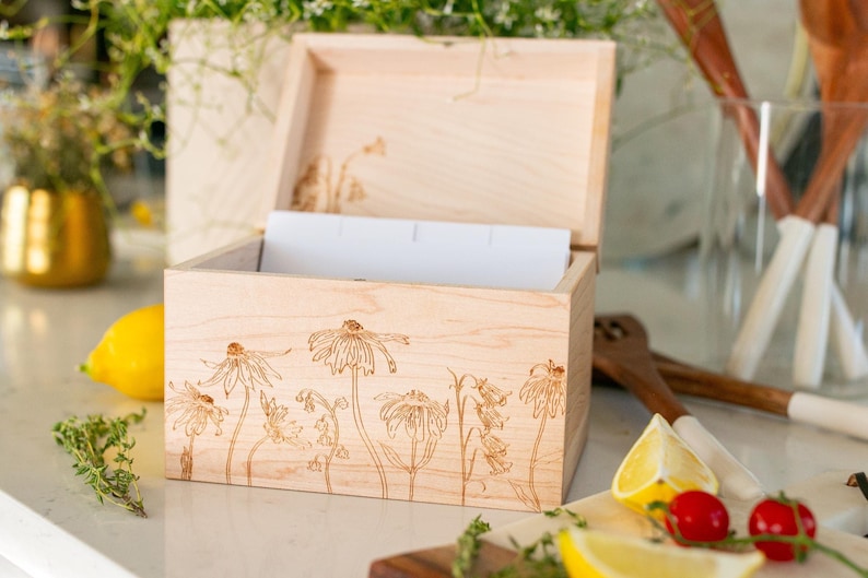 Wooden recipe box sitting with the lid open on a kitchen counter. The recipe box displays floral etching on the front of the recipe box and under the lid. Sitting inside of the box are white divider cards.