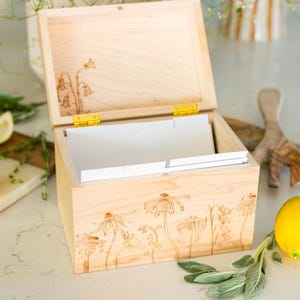 Light wooden recipe box sits on a kitchen counter with the lid halfway open. On the front of the box and under the lid of the box is a floral design etched into the wood. Inside the recipe box sits white divider cards.