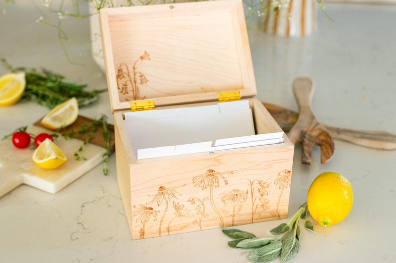 Light wooden recipe box sits on a kitchen counter with the lid halfway open. On the front of the box and under the lid of the box is a floral design etched into the wood. Inside the recipe box sits white divider cards.