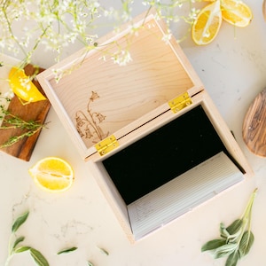 Light colored wood recipe box sitting on kitchen counter. A overhead view shows the inside of the recipe box. A black velvet lines the bottom of the box. Recipe cards and dividers sit to the side of the inside of the box.