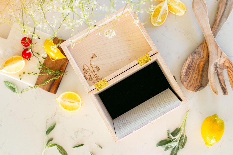 Light colored wood recipe box sitting on kitchen counter. A overhead view shows the inside of the recipe box. A black velvet lines the bottom of the box. Recipe cards and dividers sit to the side of the inside of the box.