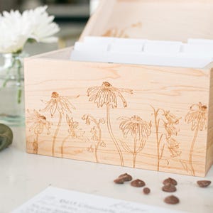 Recipe box made of light colored wood sits on kitchen counter. The lid is open but cut off in the photo as the main focus of the image displays the floral etching on the front of the recipe box.