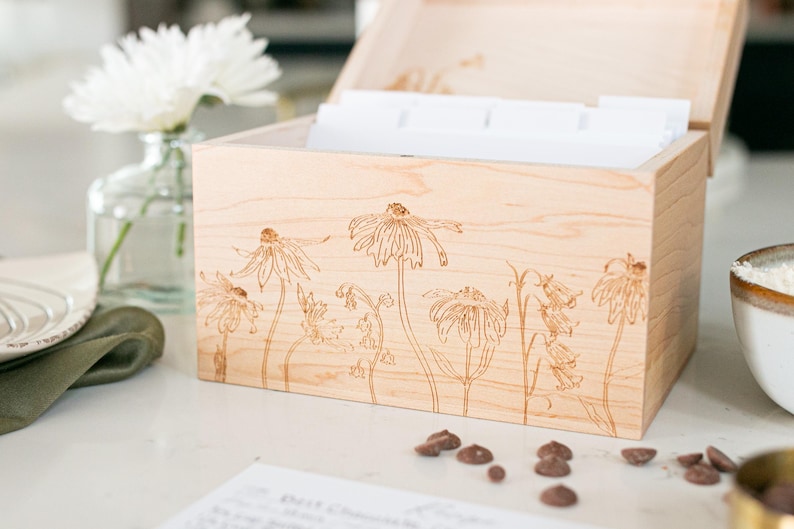Recipe box made of light colored wood sits on kitchen counter. The lid is open but cut off in the photo as the main focus of the image displays the floral etching on the front of the recipe box.