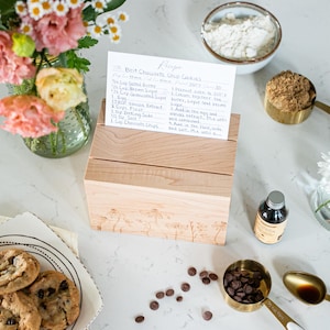 Wooden recipe box sits on a kitchen counter. The lid of the recipe box is closed. On top of the recipe box are two notches. A recipe card is propped up in one of the notches displaying ingredients and cooking instructions.