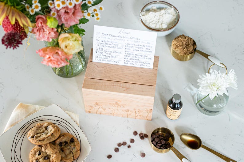 Wooden recipe box sits on a kitchen counter. The lid of the recipe box is closed. On top of the recipe box are two notches. A recipe card is propped up in one of the notches displaying ingredients and cooking instructions.