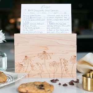 Light Colored wood recipe box sits on a kitchen counter. The front of the recipe box has a beautiful floral design etched into the wood. A recipe card with chocolate chip cookie ingredients is propped on top of the lid of the box.