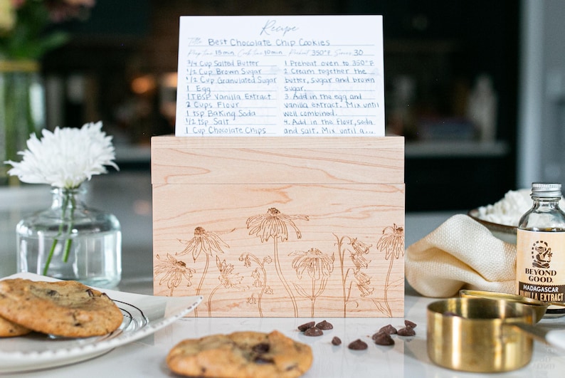 Light Colored wood recipe box sits on a kitchen counter. The front of the recipe box has a beautiful floral design etched into the wood. A recipe card with chocolate chip cookie ingredients is propped on top of the lid of the box.