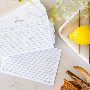 A stack of 50 gray and white recipe cards sits on a white marble kitchen counter. Recipe cards have a light beautiful floral design. One of the cards sits next to the stack and shows the design of the back of the card (college ruled and flowers).