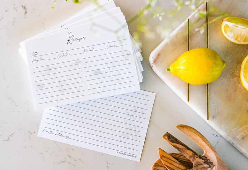 A stack of 50 gray and white recipe cards sits on a white marble kitchen counter. Recipe cards have a light beautiful floral design. One of the cards sits next to the stack and shows the design of the back of the card (college ruled and flowers).