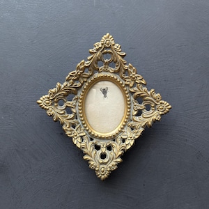 May include: A diamond-shaped picture frame with an ornate gold finish and intricate floral scrollwork. The frame features a central oval opening containing a cream-colored insert with a small, black, detailed illustration of a bee at the top.