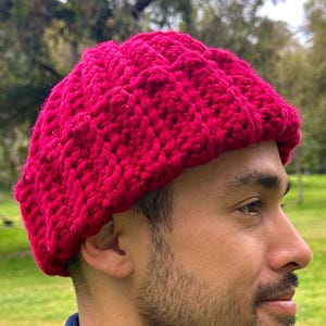 May include: A vibrant red crocheted hat. The hat is a deep, rich shade of red and appears to be handmade. The hat is worn by a person wearing a blue jacket. The background is a park with green grass and trees.