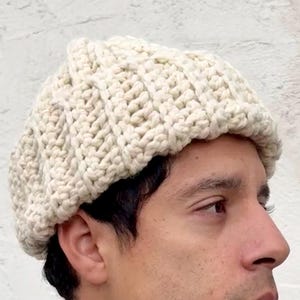 May include: Cream-colored, hand-crocheted beret. The hat is made of thick yarn, with a ribbed texture. The beret is worn by a person. The person is wearing a striped shirt and a purple t-shirt.