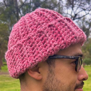 May include: A pink, hand-knitted beanie hat. The hat is made of a chunky, textured yarn and has a ribbed brim. The hat is worn by a person wearing sunglasses and a blue jacket. The background is a park with trees.