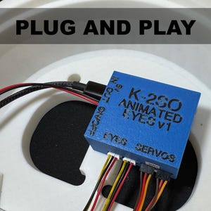 Puede incluir: Módulo azul K-2SO Animated Eyes V1 con múltiples cables de colores y el texto "EYES" y "SERVOS". El módulo está conectado a un cable negro y se encuentra sobre un fondo blanco con el texto "PLUG AND PLAY".