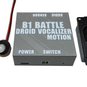 Könnte beinhalten: Ein graues, rechteckiges elektronisches Steuergehäuse mit der Aufschrift B1 BATTLE DROID VOCALIZER MOTION sowie den Texten POWER, SWITCH, AUDIO und SENSOR. Es enthält zwei verkabelte Komponenten mit einem schwarzen Knopf und einem weißen Sensor.