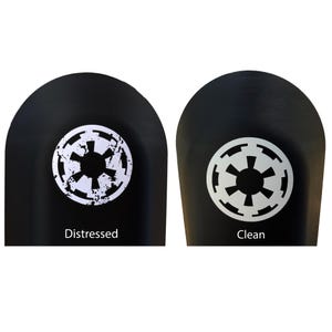 May include: Two black, rounded surfaces featuring white circular emblems. The left emblem displays a distressed, weathered version of the Imperial symbol with the text "Distressed" below it. The right emblem shows a clean, solid version with the text "Clean" below it.