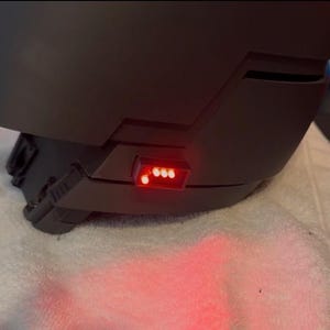 Puede incluir: Un casco gris oscuro con un panel de luz rojo rectangular con tres puntos iluminados. El casco tiene una parte superior redondeada con una gran abertura y está sobre una toalla blanca. La imagen es un primer plano.