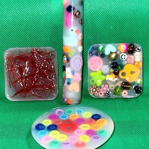 May include: Four clear resin coasters with different colorful designs. One coaster has red glitter, one has colorful beads, one has colorful rings, and one has a vertical design with colorful beads.