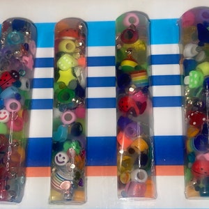 Puede incluir: Cuatro tubos de plástico transparente llenos de cuentas y abalorios de colores. Los tubos miden aproximadamente 25 cm de largo y 2,5 cm de diámetro. Las cuentas y los abalorios son de diversas formas y tamaños, incluyendo estrellas, corazones, caritas sonrientes y mariquitas.