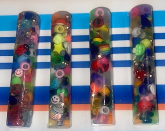 Puede incluir: Cuatro tubos de plástico transparente llenos de cuentas y abalorios de colores. Los tubos miden aproximadamente 25 cm de largo y 2,5 cm de diámetro. Las cuentas y los abalorios son de diversas formas y tamaños, incluyendo estrellas, corazones, caritas sonrientes y mariquitas.
