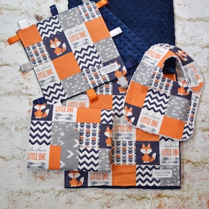 May include: A baby gift set with a blue and orange patchwork design featuring foxes, arrows, and the text "Be brave little one" and "Too cute". The set includes a burp cloth, a bib, and a soft blanket.