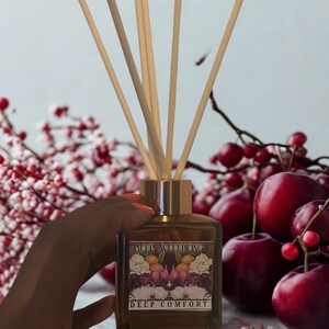 Op de afbeelding: Een bruine glazen diffuserfles met houten stokjes, met het label "DEEP COMFORT". De fles heeft een goudkleurige dop en wordt vastgehouden voor een achtergrond van rode bessen en roze bloemen. Het label heeft een bloemenontwerp.