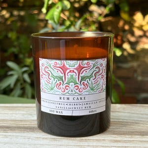 May include: A brown glass candle jar with a label that reads "RUM CAKE" and lists the ingredients: "ORANGE, NUTMEG, CHERRY, JAMAICAN SPICE, VANILLA, SWEET RUM". The label also states "SOY WAX" and "40hrs".