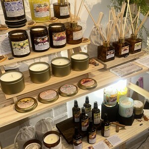 K&ouml;nnte beinhalten: Ausstellung verschiedener Duftkerzen, Schilf-Diffusoren und &auml;therischer &Ouml;le auf Holzregalen. Die Kerzen befinden sich in Gl&auml;sern und Metalldosen, mit Etiketten, die D&uuml;fte wie Grapefruit und Patchouli angeben. Schilf-Diffusoren in braunen Flaschen.