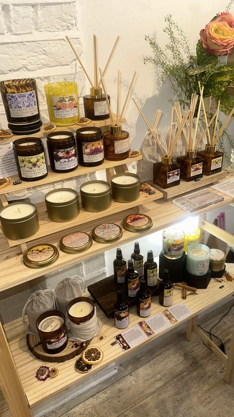 K&ouml;nnte beinhalten: Ausstellung verschiedener Duftkerzen, Schilf-Diffusoren und &auml;therischer &Ouml;le auf Holzregalen. Die Kerzen befinden sich in Gl&auml;sern und Metalldosen, mit Etiketten, die D&uuml;fte wie Grapefruit und Patchouli angeben. Schilf-Diffusoren in braunen Flaschen.