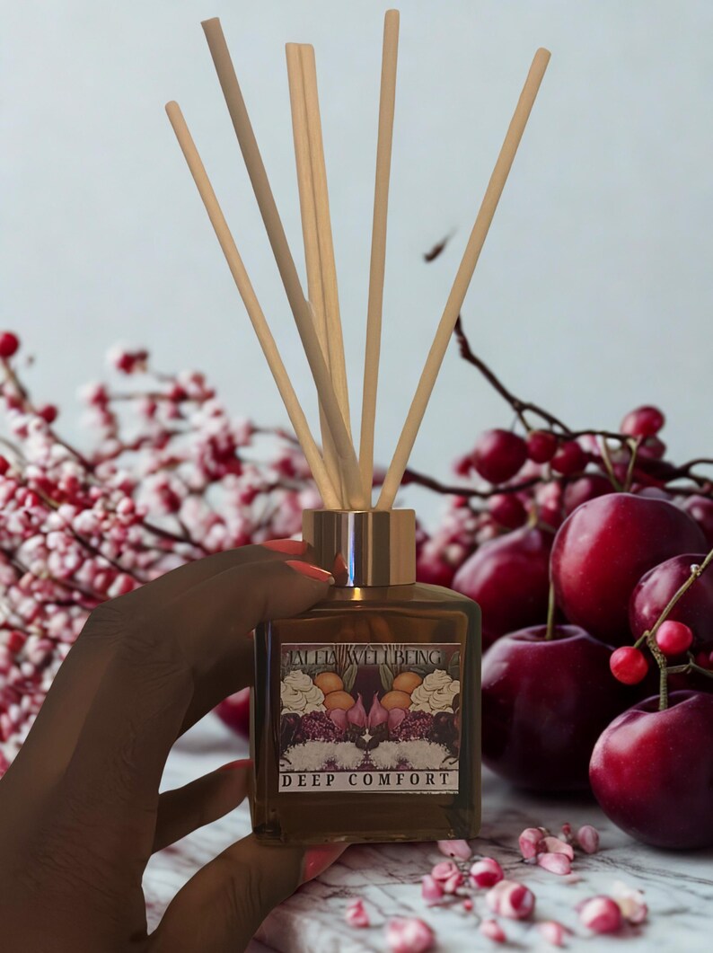 K&ouml;nnte beinhalten: Eine braune Glas-Diffusorflasche mit Holzst&auml;bchen, beschriftet mit "DEEP COMFORT". Die Flasche hat einen goldfarbenen Deckel und wird vor einem Hintergrund aus roten Beeren und rosa Bl&uuml;ten gehalten. Das Etikett hat ein florales Design.