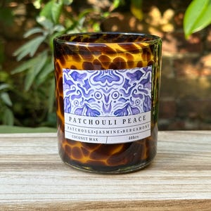 Puede incluir: Una vela marrón y beige con un patrón de caparazón de tortuga. La vela está etiquetada como "Patchouli Peace" y tiene las notas de aroma "Patchouli, Jazmín, Bergamota". La vela está hecha de cera de coco y tiene un tiempo de combustión de 40 horas.