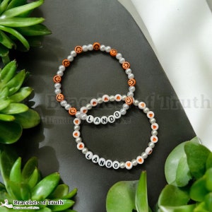 Op de afbeelding: Twee kralenarmbanden met witte kralen en oranje smiley- en hartaanhangers. Een armband spelt de naam "LANDO" in witte kralen.