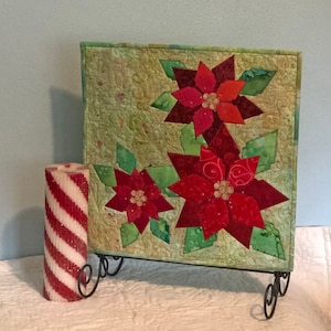 May include: A green fabric wall hanging with three red poinsettia flowers. The wall hanging is on a black metal stand. A red and white striped candle is in the foreground.