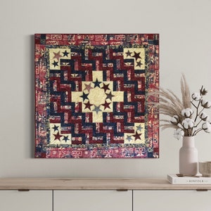 May include: A red, white, and blue quilt with a star pattern. The quilt is hanging on a white wall above a wooden shelf with a book and a vase of cotton flowers.