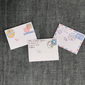K&ouml;nnte beinhalten: Drei wei&szlig;e Umschl&auml;ge mit handgezeichneten Illustrationen von Feen, einem Zahn und einem Pilz. Die Umschl&auml;ge sind an "An :" adressiert und haben Briefmarken mit den Worten "Fairy Mail" und "Fairy Post Service".