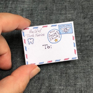 K&ouml;nnte beinhalten: Ein wei&szlig;er Umschlag mit roten und blauen Streifen. Der Umschlag hat eine Zeichnung eines Zahns und den Text "Guild of Tooth Fairies". Au&szlig;erdem hat er eine Briefmarke mit einer Fee und dem Text "Fairy Post Office" und "Fairy Mail". Der Umschlag ist adressiert mit "To:"