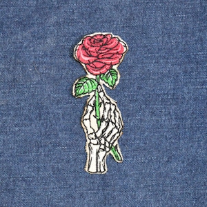 May include: A skeleton hand holding a red rose with green leaves. The rose is embroidered on a white fabric patch.