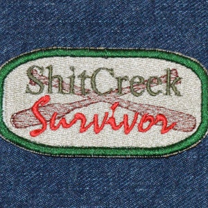 Può includere: Una toppa ovale bianca con bordo verde e il testo "Shit Creek Survivor" in ricamo marrone e rosso. Due pagaie di legno incrociate sono ricamate in marrone.