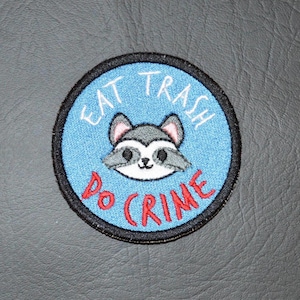 May include: Embroidered patch with a raccoon illustration inside a blue circle. The text "EAT TRASH DO CRIME" is embroidered in white and red. The patch has a black border.