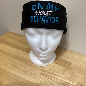 Può includere: Fascia nera con la scritta ricamata "ON MY worst BEHAVIOR" in blu e bianco. La fascia è esposta su una testa di manichino bianca, su una superficie in legno chiaro.
