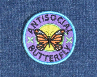 Könnte beinhalten: Gestickter Aufnäher mit den Worten "ANTISOCIAL BUTTERFLY" in Schwarz um einen lila Kreis. Der Aufnäher zeigt einen Schmetterling mit orangefarbenen und schwarzen Flügeln auf gelbem Grund, mit grünen Sternakzenten.