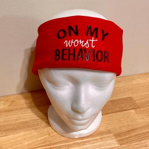 Può includere: Una fascia rossa con la scritta "ON MY worst BEHAVIOR" in bianco e nero. La fascia è esposta su una testa di manichino in schiuma bianca, su sfondo chiaro.