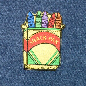 May include: A yellow and red embroidered patch of a crayon box with the text "SNACK PAK" on the front. The box is filled with six crayons in various colors.