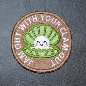May include: Round embroidered patch with a brown border and the text "JAM OUT WITH YOUR CLAM OUT" in white. The patch features a green clam shell with a smiling pearl inside.