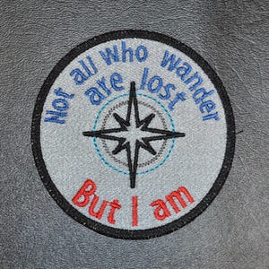 Puede incluir: Un parche redondo bordado de color gris con un diseño de rosa de los vientos y el texto "Not all who wander are lost, but I am".