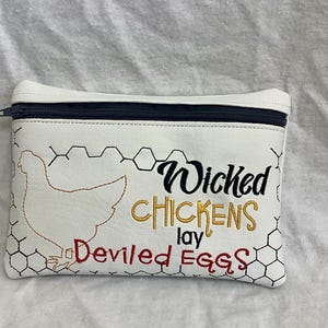 May include: White zippered pouch with a black zipper and the embroidered phrase "Wicked Chickens lay Deviled Eggs." The design includes a chicken outline and honeycomb pattern.