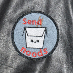 May include: A black and white embroidered patch with a blue background. The patch features a cartoon takeout container with chopsticks and the text "Send Noods".