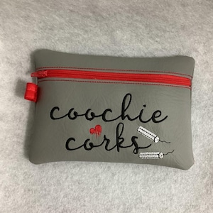 May include: A grey zipped pouch with a red zipper. The pouch has the text "coochie corks" embroidered on it in black thread. There is a red heart with a red drip above the word "corks". Two white tampons are embroidered on the pouch.