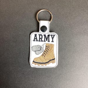 Może przedstawiać: Biały brelok do kluczy z brązowym butem, dwie srebrne identyfikatory dla psa i wyszytym czarnym napisem "ARMY".