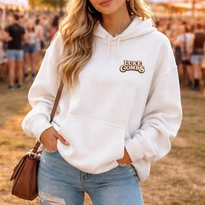 Puede incluir: Una sudadera con capucha blanca con el texto "LUKE COMBS" en letras marrones y beige. La sudadera tiene un bolsillo delantero y se usa con una gorra de béisbol y vaqueros azul claro. También se ve un bolso de hombro marrón.